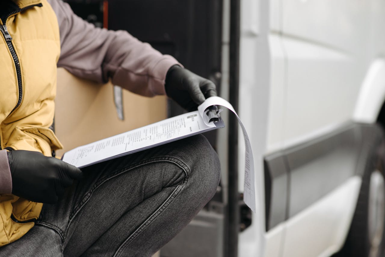 A delivery worker in gloves checking documents on a clipboard by a van.